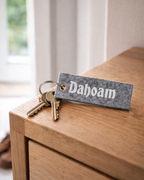 Handgemachter Schlüsselanhänger aus grauem Filz mit der Aufschrift Dahoam auf einem Schlüsselbund, welcher auf einem Sideboard liegt. 