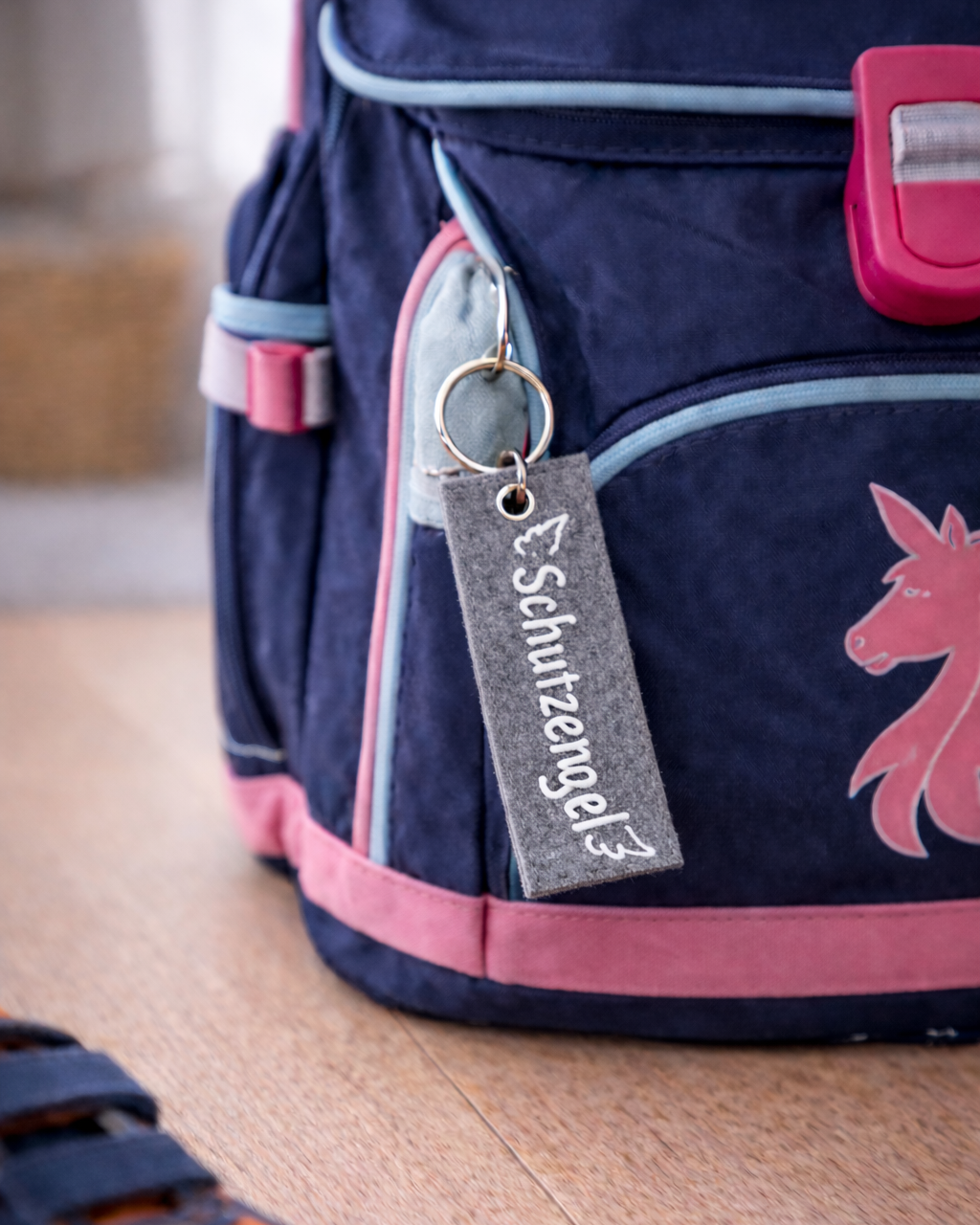 Handgemachter Schlüsselanhänger aus grauem Filz mit der Aufschrift Schutzengel auf einer Schultasche. 